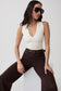 A woman wearing a fitted off-white sleeveless top with a deep V-neck, dark brown wide-leg pants, and dark sunglasses, posing on a white block against a light gray studio background.