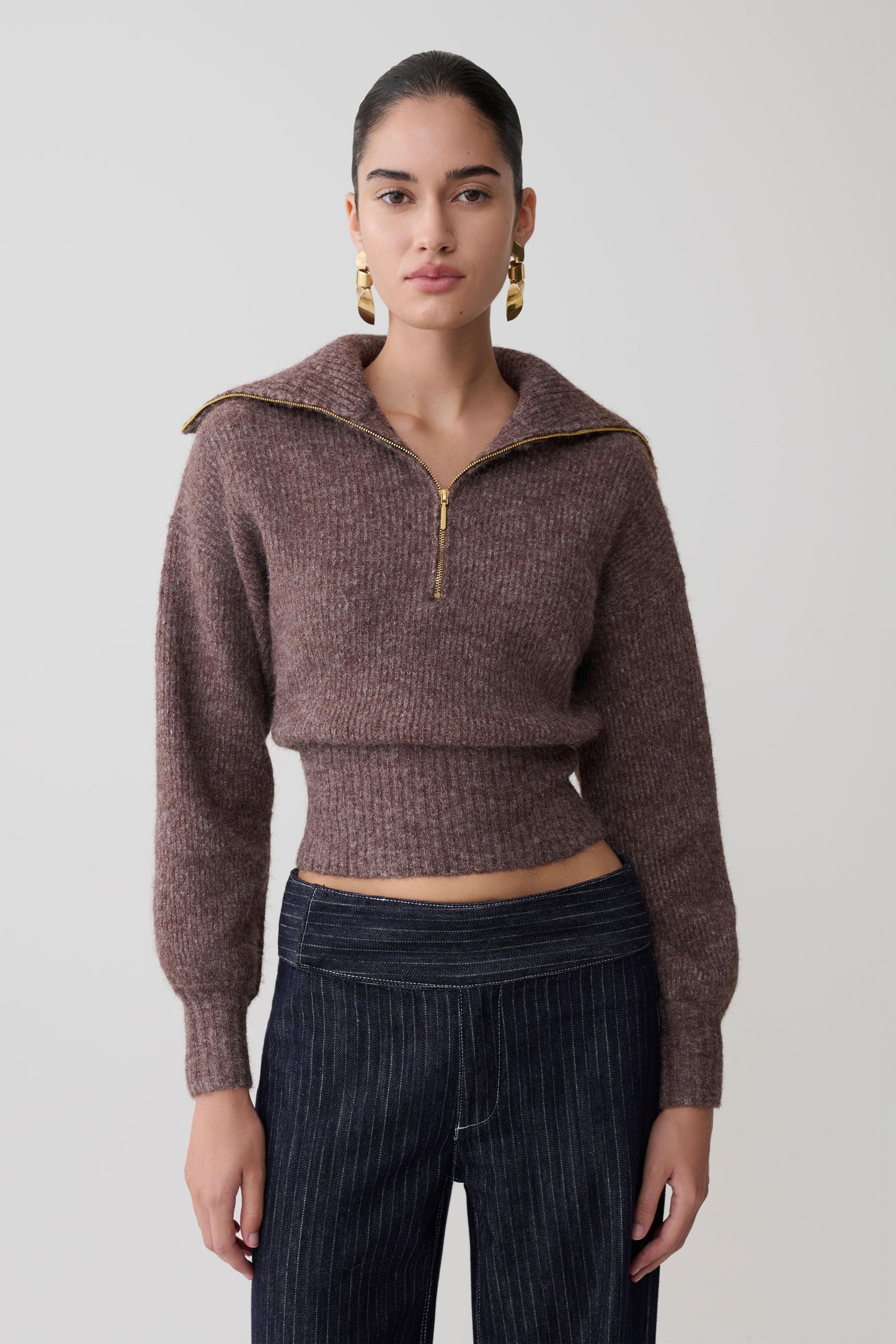 Woman wearing a brown cropped sweater with a high collar and zipper on a plain background.