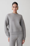 Woman wearing a gray sweater and pants on a plain background.