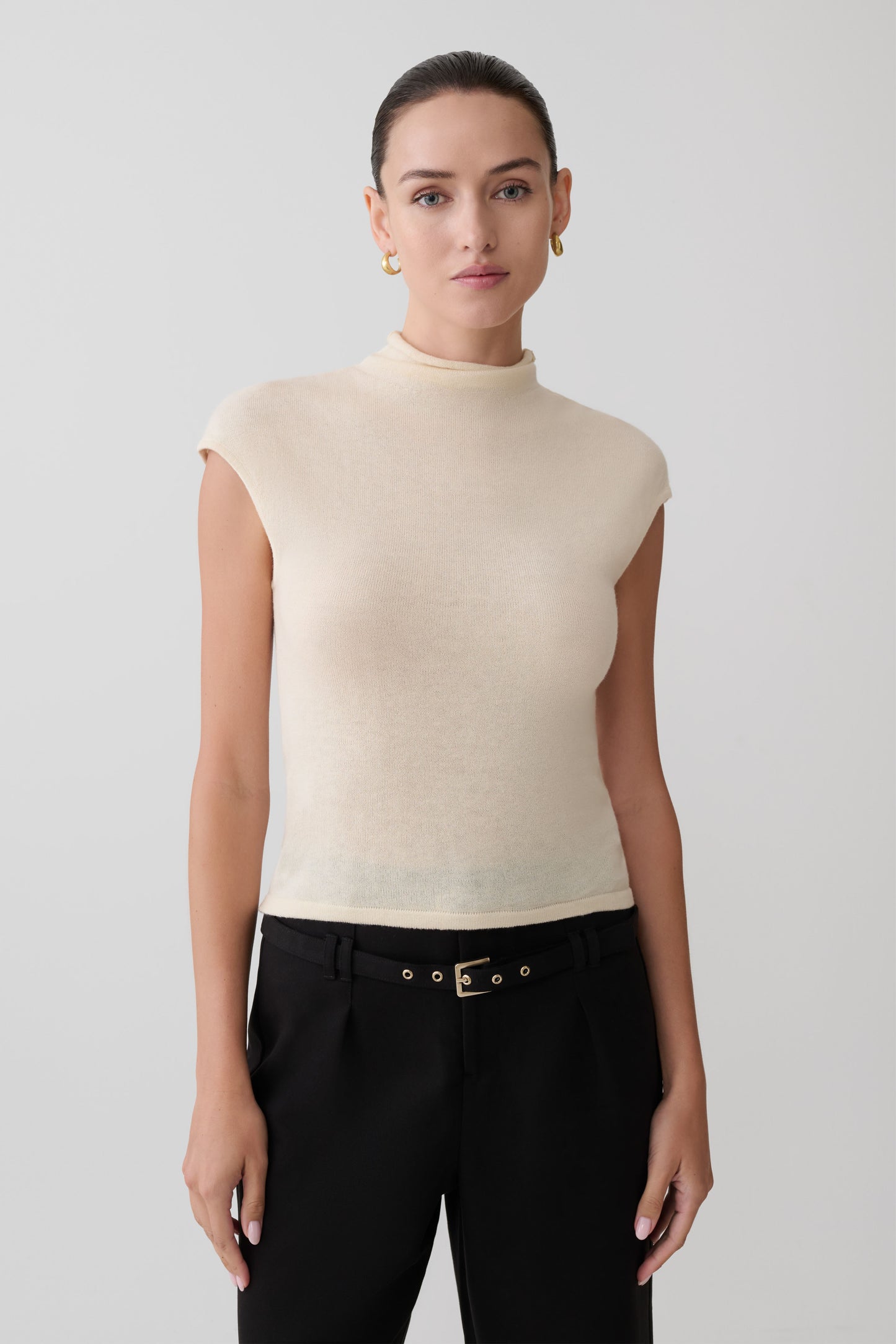 Woman wearing a beige sleeveless sweater and black pants against a plain background
