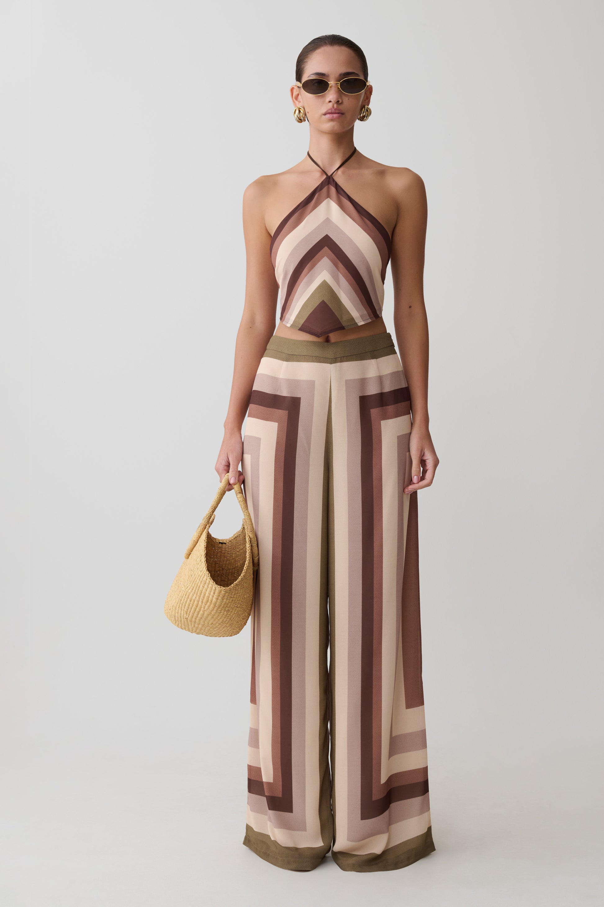 Woman wearing a patterned jumpsuit holding a straw bag on a plain background