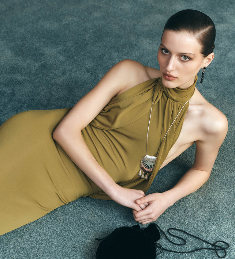 A model wearing a sleeveless olive green halter-neck dress lies on a textured teal carpet, wearing large silver statement earrings.