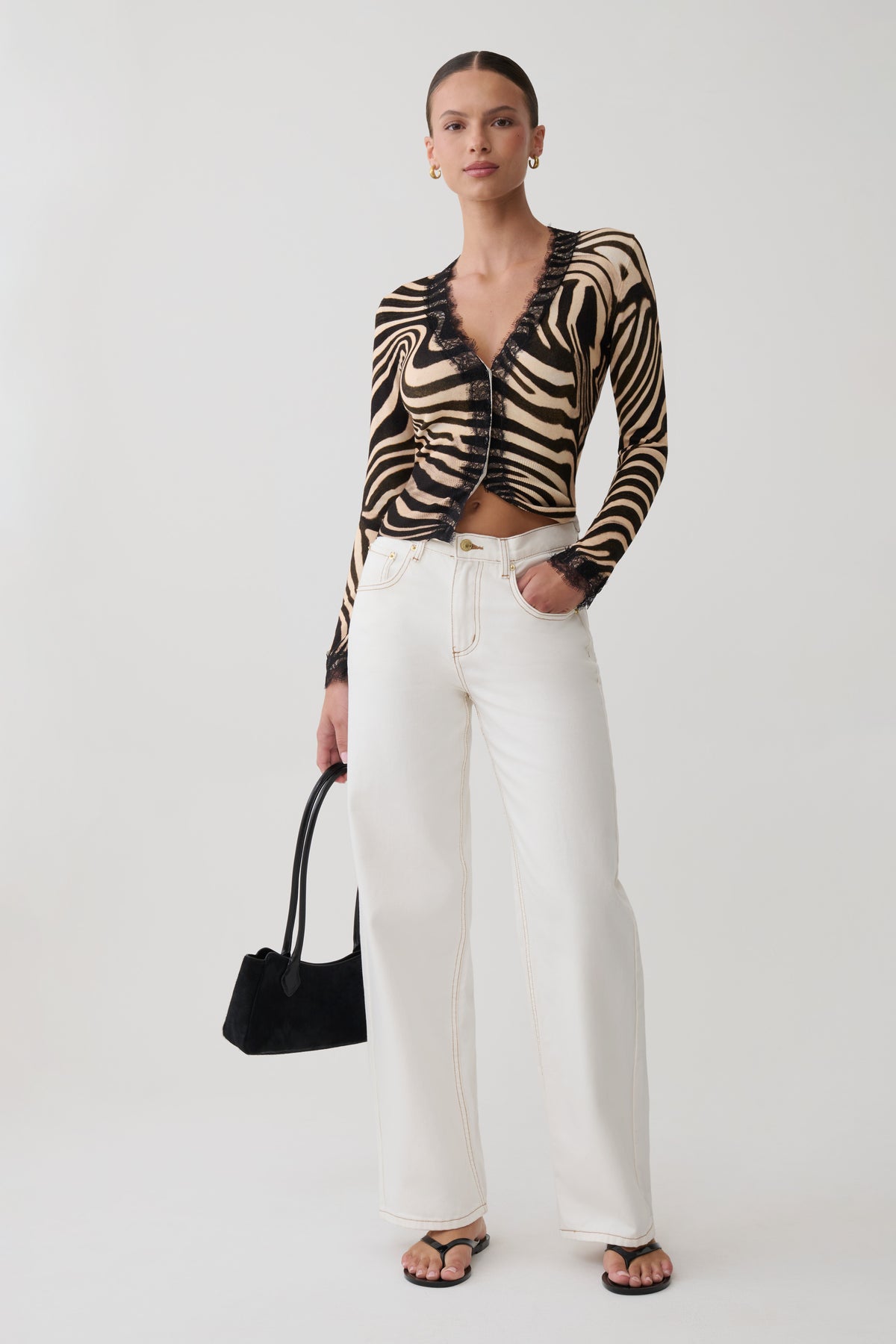 A model stands against a light gray studio background wearing a black and beige zebra-print long-sleeve crop top with ruffle trim, white wide-leg pants, and black sandals, while holding a black handbag.
