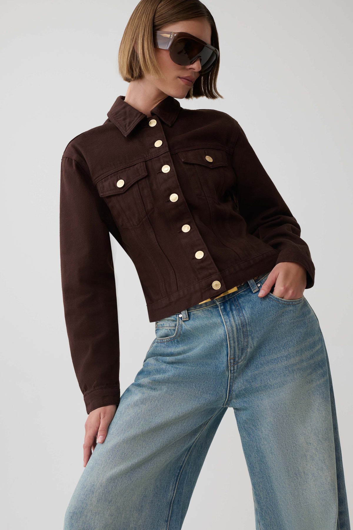 A model wearing a dark brown cropped denim jacket with gold-tone buttons and light-wash blue jeans, styled with sunglasses, against a plain light gray background.