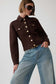 A model wearing a dark brown cropped denim jacket with gold-tone buttons and light-wash blue jeans, styled with sunglasses, against a plain light gray background.