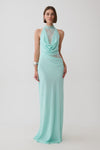A woman modeling a sleeveless mint green maxi dress with a high mock neck, draped cowl neckline, sheer lace inserts at the bust and waist, and a flowing skirt, photographed against a light gray studio background.