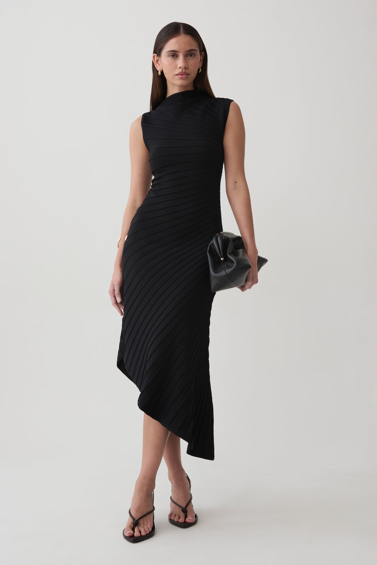 A woman modeling a sleeveless black pleated midi dress with an asymmetrical hem, paired with black strappy sandals and a small black handbag, against a plain light gray studio background.