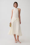 A model wearing a sleeveless cream-colored halter midi dress with a textured knit bodice and a flowing pleated skirt, styled with a small woven clutch and white sandals against a light gray studio background.