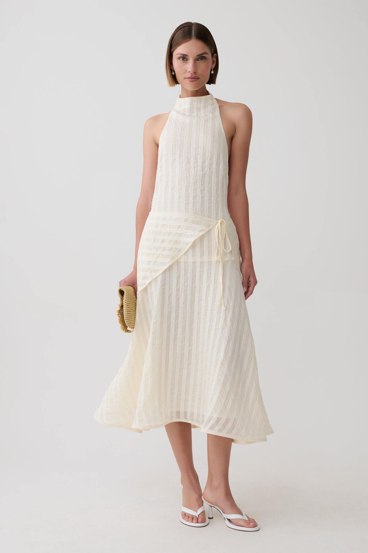 A model wearing a sleeveless cream-colored halter midi dress with a textured knit bodice and a flowing pleated skirt, styled with a small woven clutch and white sandals against a light gray studio background.