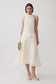 A model wearing a sleeveless cream-colored halter midi dress with a textured knit bodice and a flowing pleated skirt, styled with a small woven clutch and white sandals against a light gray studio background.