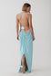 A woman modeling a light blue maxi dress from the back, showing an open back with thin straps and a draped skirt, against a plain light gray studio background.