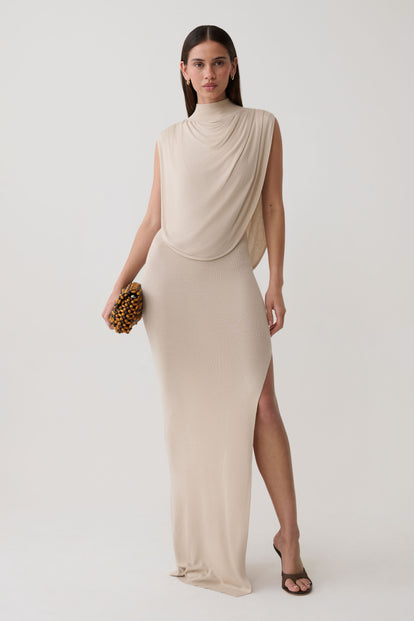 A woman models a sleeveless, draped high-neck maxi dress in a light beige/cream color, holding a small patterned clutch against a plain light gray studio background.