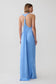 A woman shown from the back wearing a light blue satin maxi dress with a halter-style open back and a prominent gold O-ring detail at the neckline, set against a plain light gray studio background.