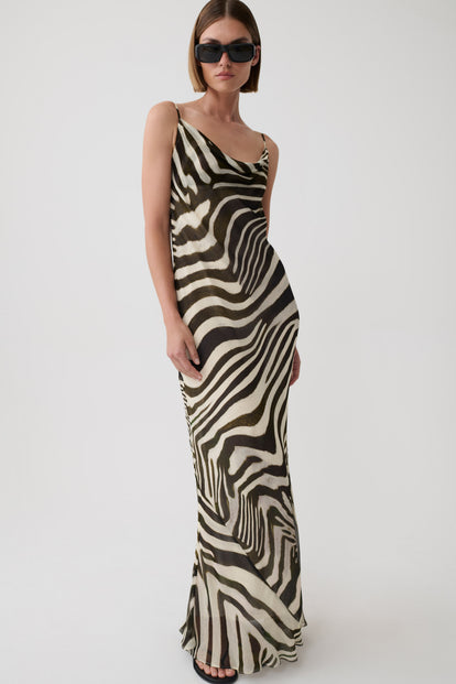 A woman modeling a sleeveless, floor-length zebra-print dress with a draped neckline and thin straps, photographed against a plain light gray studio background.