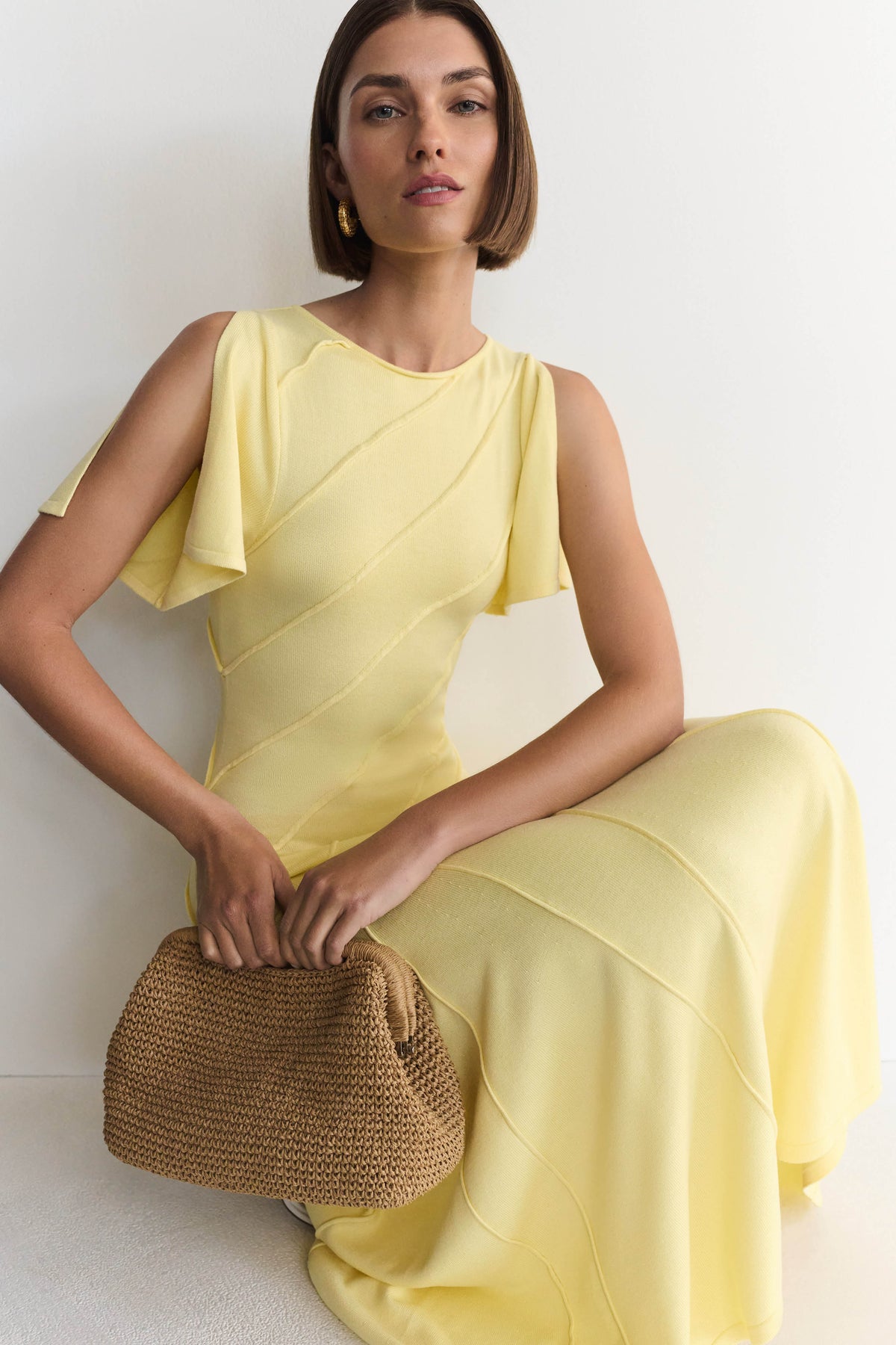 A woman wearing a pale yellow draped sleeveless dress sits against a white wall, holding a woven straw clutch bag.