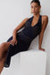 A woman models a sleeveless dark navy midi dress with textured geometric paneling, leaning against a white block on a light gray studio background.