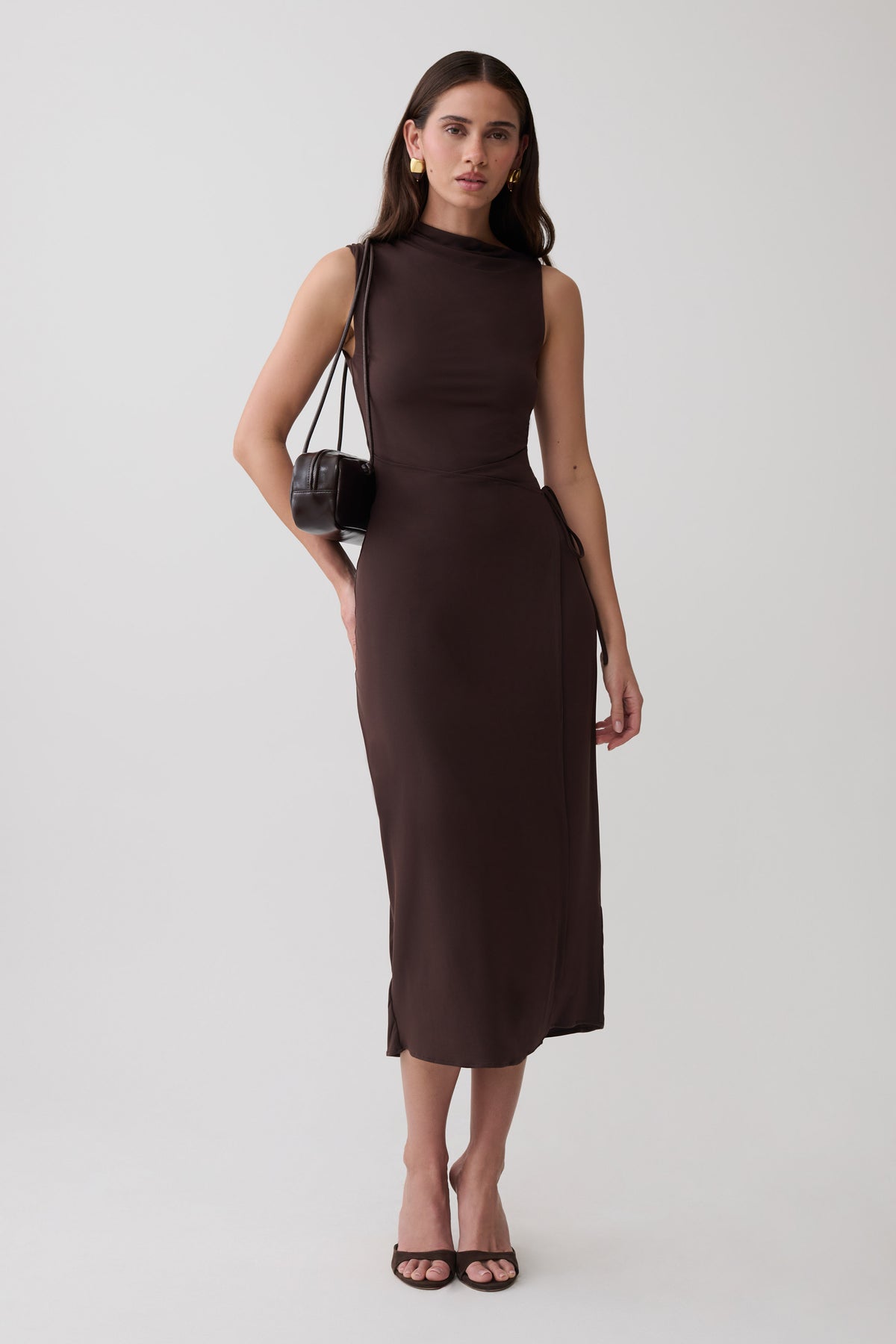 A woman modeling a sleeveless, high-neck, knee-length dark brown dress against a plain light gray studio background. She is carrying a small brown shoulder bag and wearing black heeled sandals.