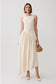 A woman models a sleeveless cream-colored midi dress with a structured bodice and a flowing skirt, holding a small woven straw handbag against a light gray studio background.