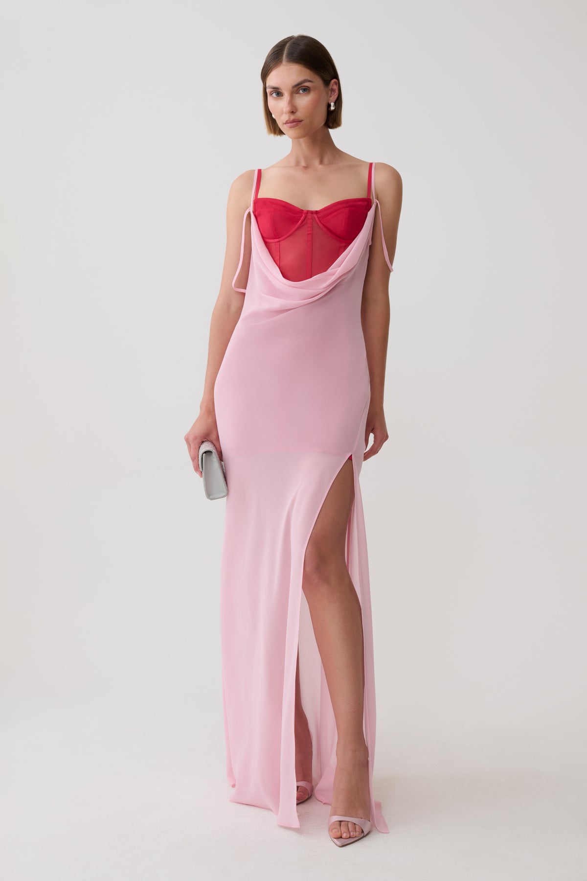 A model wearing a light pink slip-style maxi dress with a sweetheart neckline, sheer mesh bust, and a high thigh slit, paired with a bright red mesh corset mini dress underlayer and holding a small silver clutch against a plain light gray studio background.