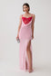 A model wearing a light pink slip-style maxi dress with a sweetheart neckline, sheer mesh bust, and a high thigh slit, paired with a bright red mesh corset mini dress underlayer and holding a small silver clutch against a plain light gray studio background.
