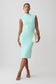 A woman modeling a sleeveless, high-neck, knee-length bodycon dress in a light mint green color, paired with a cream clutch and white strappy heels, against a light gray studio background.