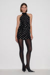 A model stands against a plain light gray studio background wearing a sleeveless black halter mini dress covered in small silver studs, sheer black tights, and black pointed heels.