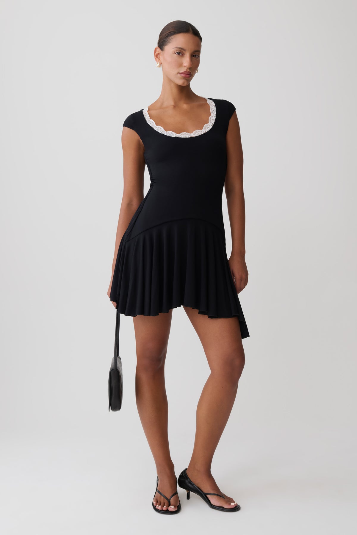 A woman modeling a black sleeveless mini dress with a white scalloped neckline, black strappy heels, and holding a black handbag against a light gray studio background.