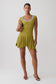 A model wearing a short olive green dress with a ruffled hem and a light blue scalloped scoop neckline, holding a small gold clutch against a light gray studio background.