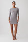 A woman modeling a fitted, three-quarter sleeve gray mini dress with a boat neckline and sheer lace trim at the hem, paired with white strappy sandals, against a plain light gray studio background.