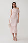A woman models a light pink off-the-shoulder ribbed midi dress with a ruffled hem and side fringe detail, paired with white strappy heels and a white faux-fur clutch, against a plain light gray studio background.