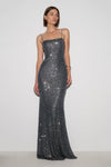 A woman modeling a floor-length, spaghetti-strap evening gown covered in dark gray sequins against a plain light gray studio background.