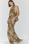 A woman modeling a sleeveless halter-neck maxi dress with a snakeskin print in beige, brown, and olive tones, featuring a high side slit and peplum waist detail, set against a plain white studio background.