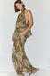 A woman modeling a sleeveless halter-neck maxi dress with a snakeskin print in beige, brown, and olive tones, featuring a high side slit and peplum waist detail, set against a plain white studio background.