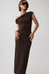 A woman modeling a dark brown, one-shoulder textured knit maxi dress with a ruched bodice and horizontal pleated skirt, paired with dark sunglasses against a light gray studio background.