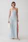 A woman models a strapless light blue pleated maxi dress with a high thigh slit, holding a white feather clutch against a plain light gray studio background.