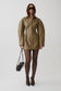 A model wearing a structured, oversized olive-brown mini jacket with a front zipper, styled with black tights, pointed-toe heels, sunglasses, and a small black handbag against a light gray studio background.