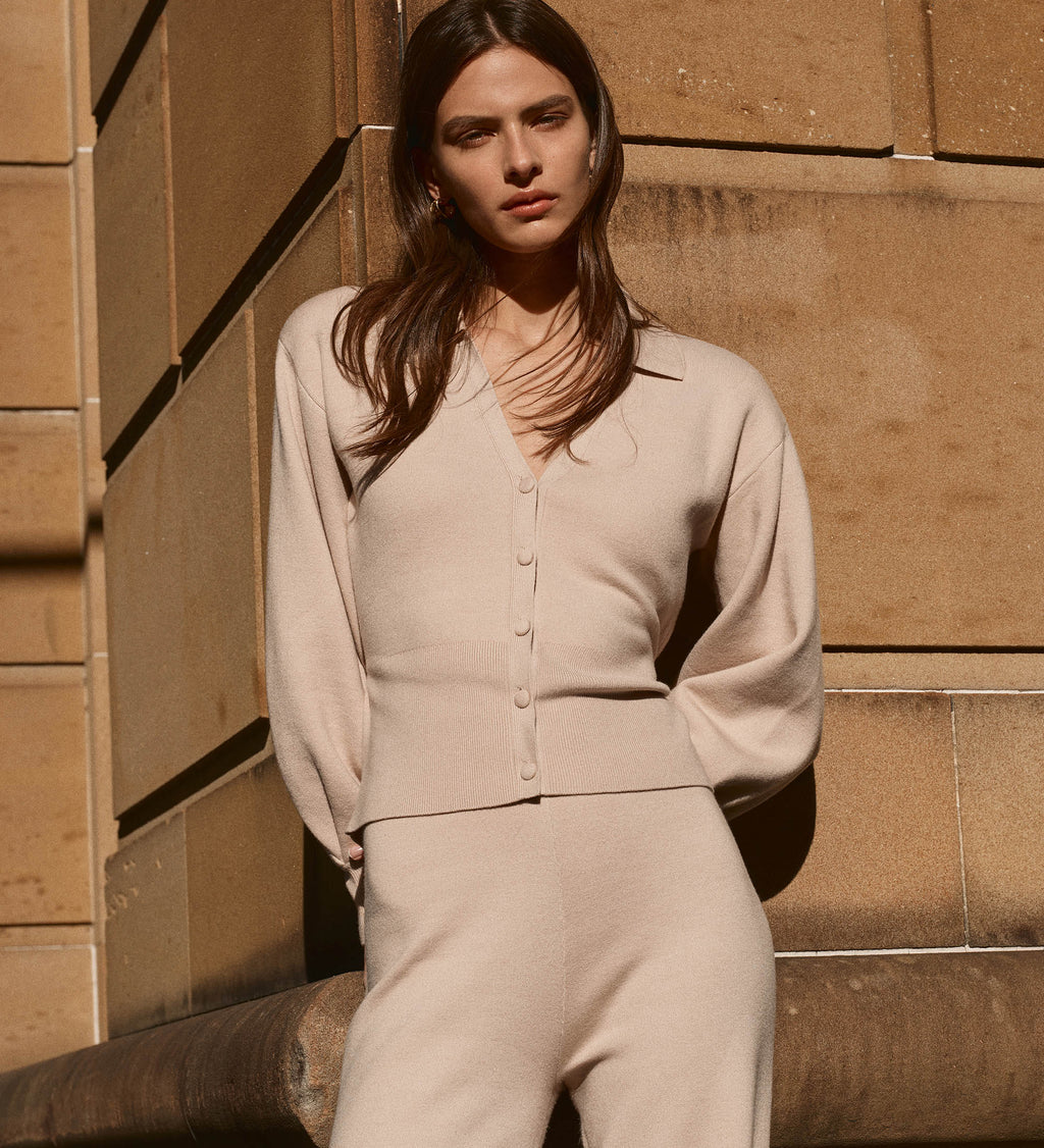 Woman in a beige outfit standing against a stone wall.