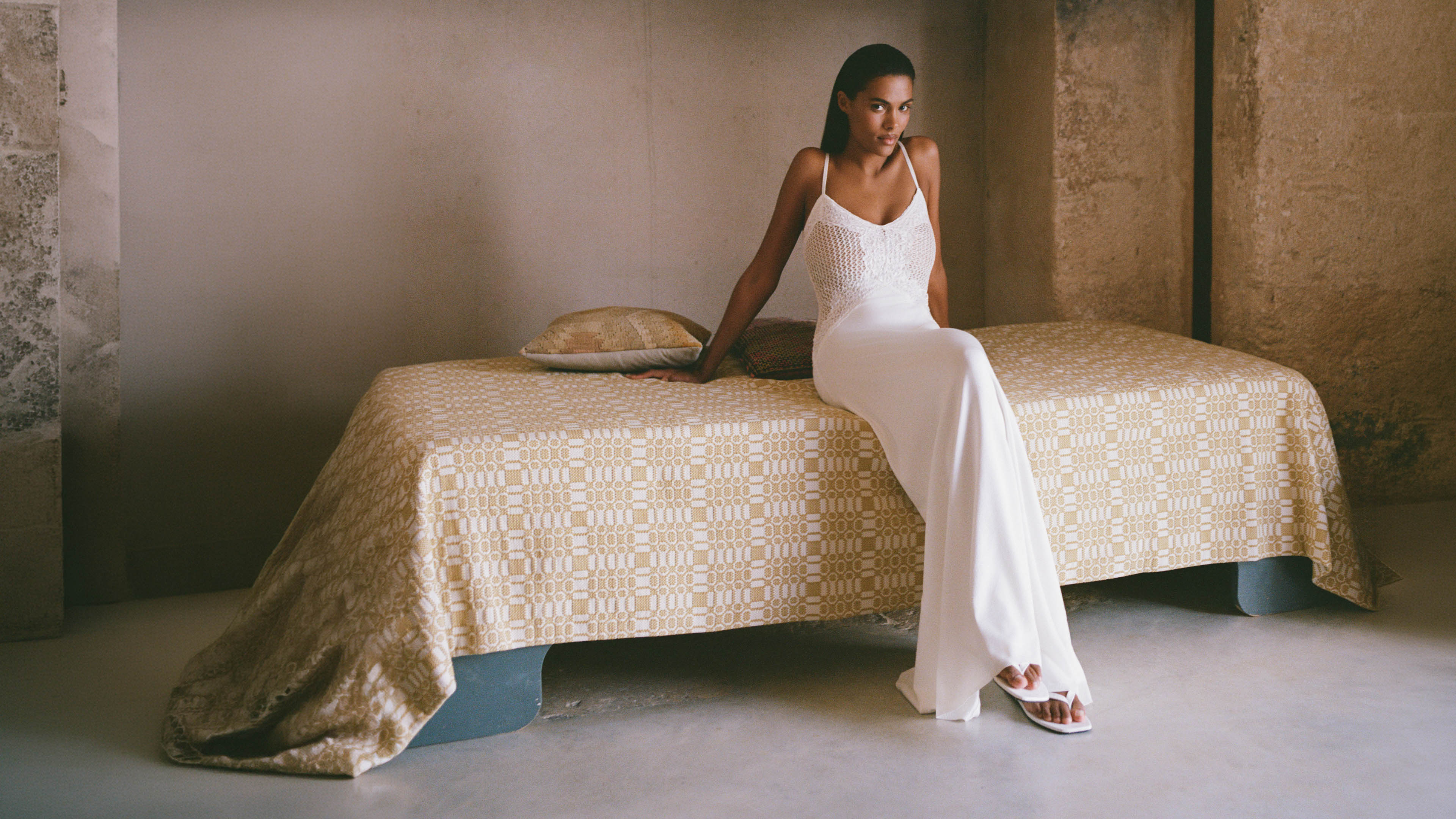 Woman in a white dress sitting on a patterned bed in a minimalistic room.