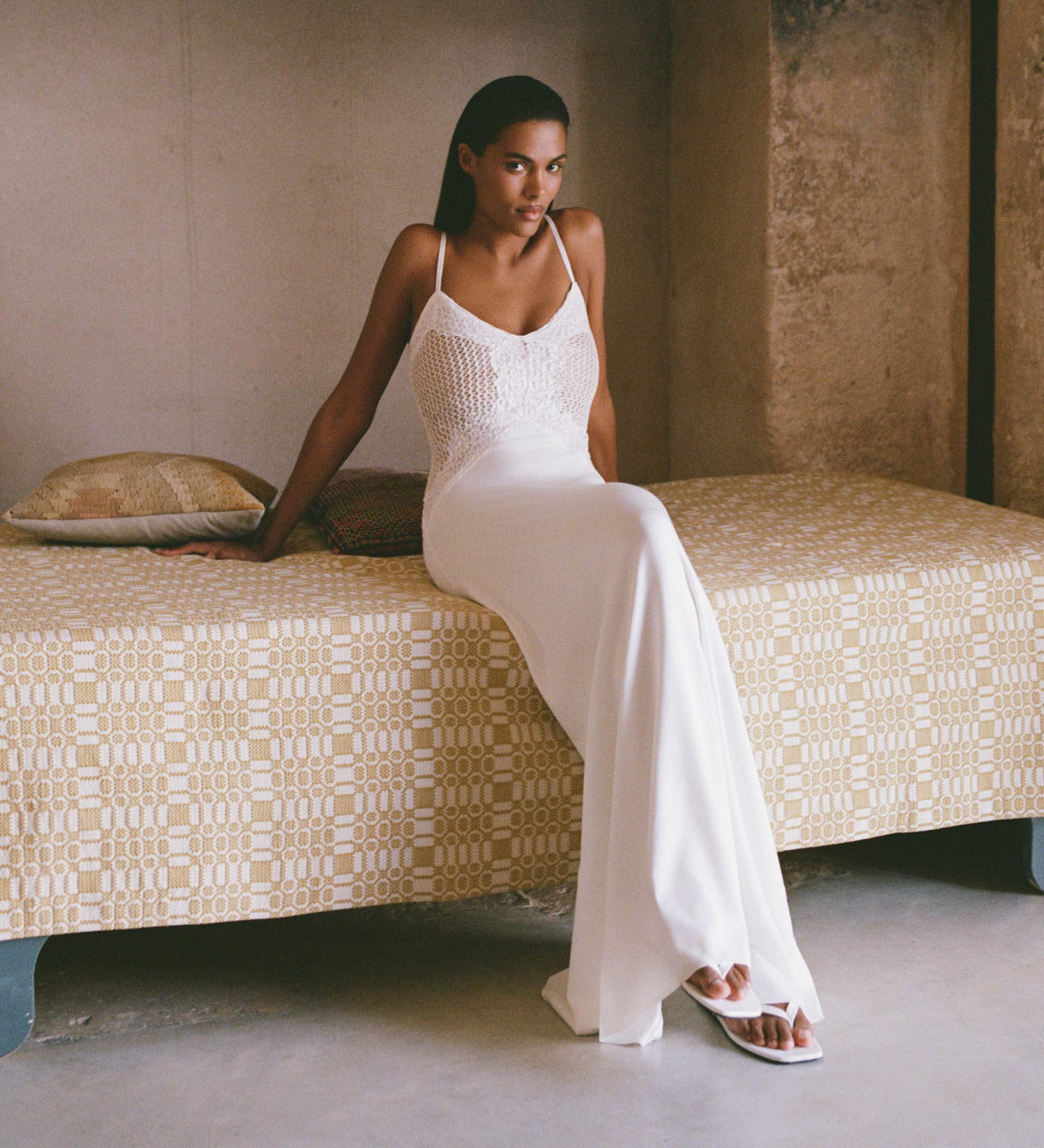 Woman in a white dress sitting on a patterned bed in a minimalistic room.