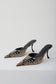 A pair of pointed-toe mule heels with a black and beige snakeskin pattern, displayed on a clean white studio background.