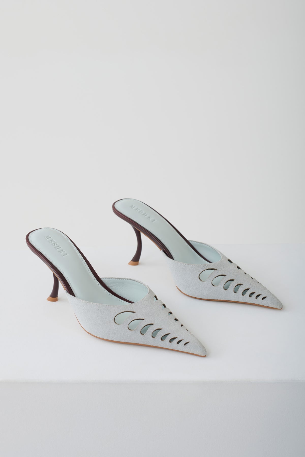 A pair of pointed-toe mule heels in a pale icy blue color, featuring decorative cutout detailing on the sides and slim dark brown stiletto heels, photographed on a clean white studio background.