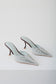 A pair of pointed-toe mule heels in a pale icy blue color, featuring decorative cutout detailing on the sides and slim dark brown stiletto heels, photographed on a clean white studio background.