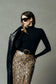 A model wearing a black long-sleeve mock neck top, a black belt with a gold buckle, a snakeskin print skirt, and black sunglasses, holding a black leather jacket against a textured gray background.