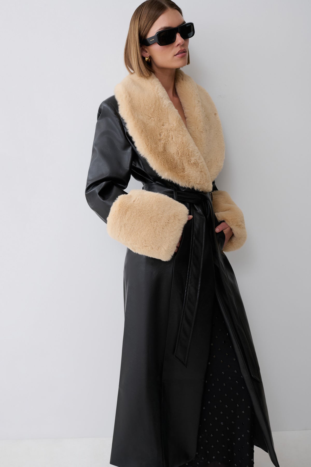 A woman modeling a long black coat with a large beige fur collar and matching beige fur cuffs, photographed against a plain light gray studio background.