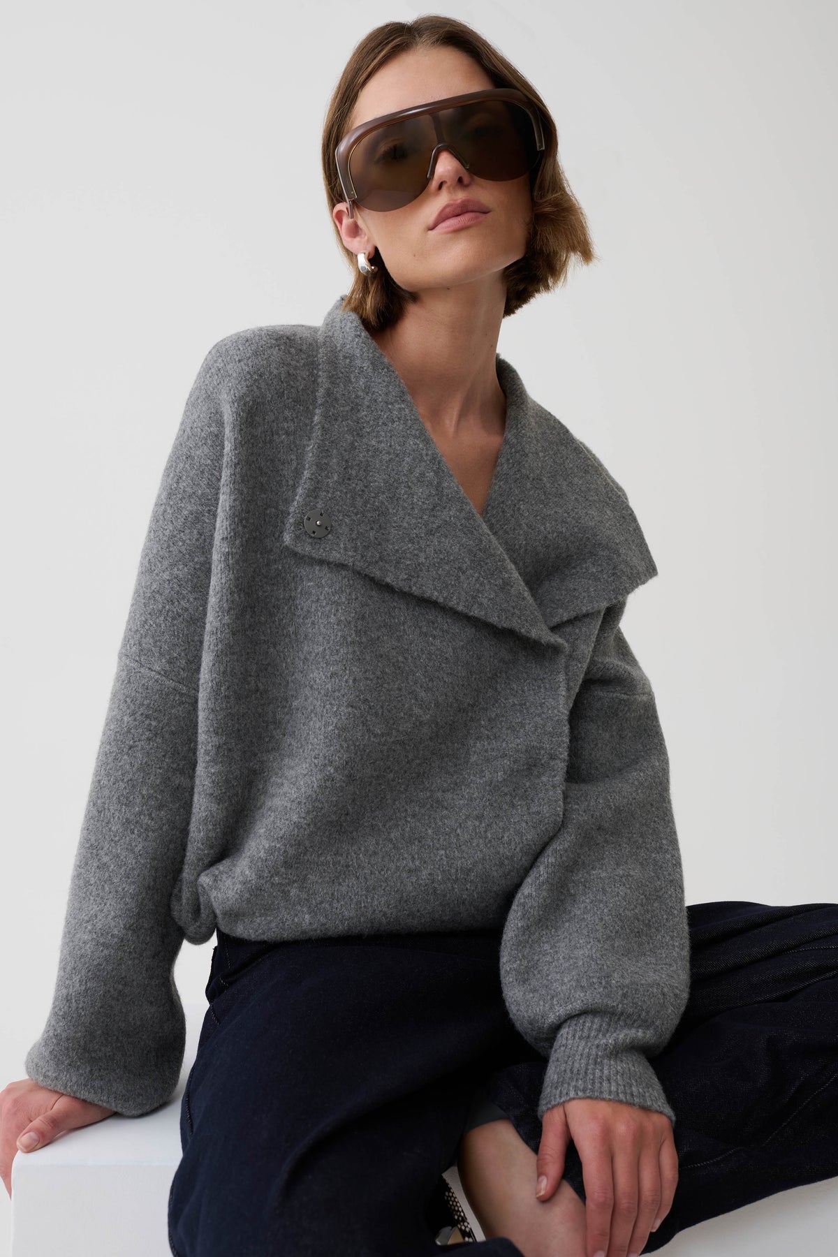 A woman wearing a gray knit wrap cardigan and dark sunglasses, seated against a plain light background.