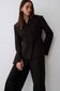 A woman modeling a black tailored blazer with structured shoulders and matching black trousers against a plain light gray studio background.