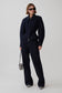 A model stands against a light gray studio background wearing a matching dark navy blue zip-up jacket and wide-leg trousers set, styled with black sunglasses, a small black-and-white zebra-patterned handbag, and black-and-white striped shoes.