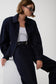 A model wearing a dark navy denim jacket over a white tank top and dark navy trousers, accessorized with a black belt and sunglasses, seated against a plain white background.