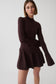 A woman modeling a dark brown ribbed knit mock-neck long-sleeve top with a matching pleated mini skirt, photographed against a light gray studio background.