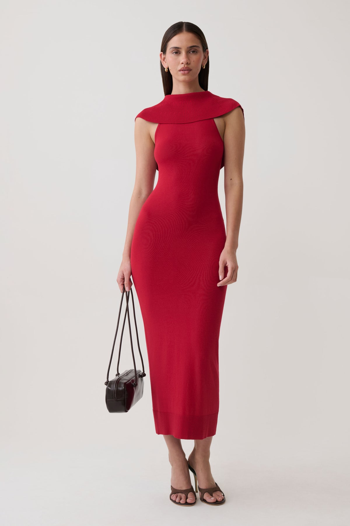 A woman modeling a fitted sleeveless red midi dress with a high mock neck and cap shoulders, holding a small black handbag against a plain light gray studio background.